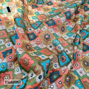 May include: A close-up of a fabric with a colorful geometric pattern. The fabric is embroidered with small mirrors and has a light beige background. The pattern features shades of pink, blue, green, and gold. The fabric is called "Embf061".