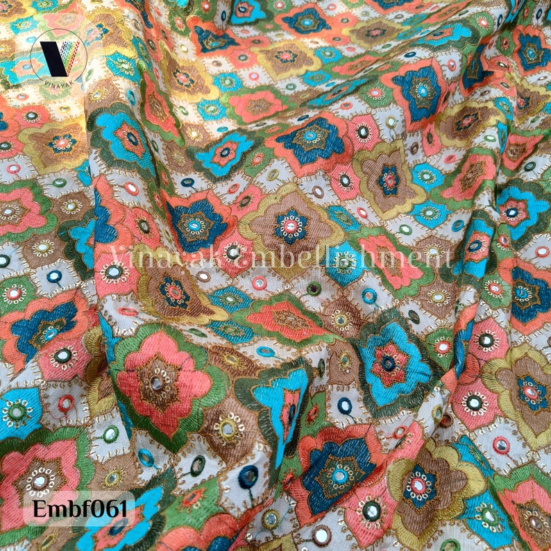 May include: A close-up of a fabric with a colorful geometric pattern. The fabric is embroidered with small mirrors and has a light beige background. The pattern features shades of pink, blue, green, and gold. The fabric is called "Embf061".