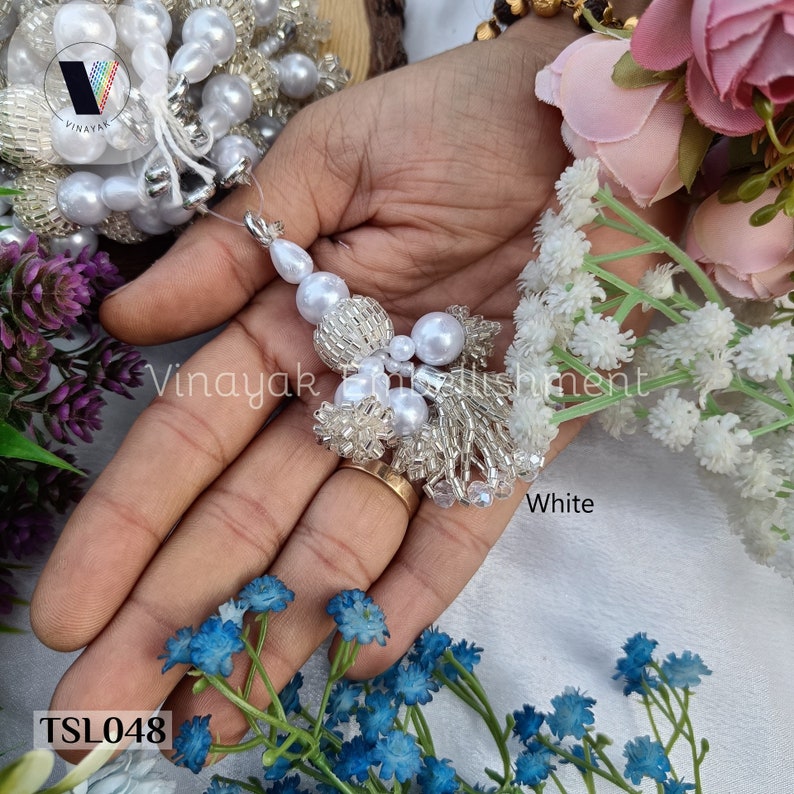 May include: White beaded tassel trim with a pearl accent. The trim is made of small white beads and pearls, and it has a tassel at the end. The trim is shown on a hand, and the text "TSL048" and "White" are visible in the image.