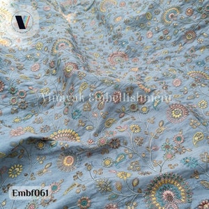 May include: A light blue fabric with a floral pattern embroidered in shades of pink, yellow, and green. The fabric has a subtle sheen and a delicate texture.
