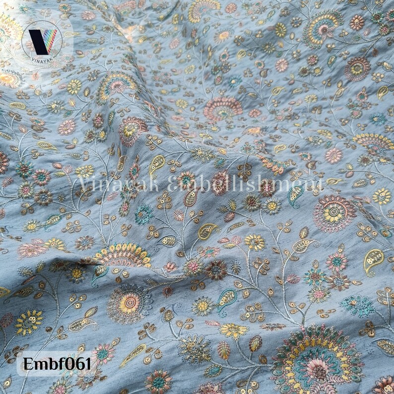 May include: A light blue fabric with a floral pattern embroidered in shades of pink, yellow, and green. The fabric has a subtle sheen and a delicate texture.