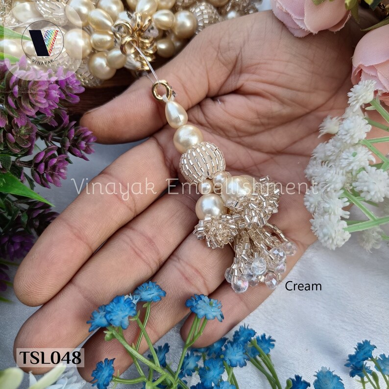 May include: Cream colored beaded tassel with a pearl and crystal embellishment. The tassel is held in a hand. The text "TSL048" and "Cream" are visible in the image.