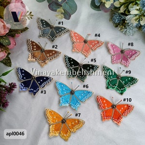 May include: A collection of ten embroidered butterfly brooches in various colors, including blue, orange, pink, and green. Each butterfly has a unique design with beaded accents and delicate antennae. The brooches are numbered and arranged on a light-colored surface.
