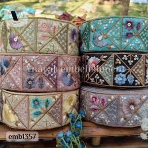 May include: Five spools of embroidered trim with floral designs in various colors, including green, blue, pink, and black. The trim is arranged in a stack, with the top spool showing a turquoise background and the bottom spool showing a light brown background. The trim is labeled "emb1357".