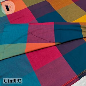 Peut inclure: Un tissu textile présentant un motif à grands carreaux dans des couleurs vives, notamment bleu sarcelle, magenta, orange, violet, vert olive et jaune. Le matériau a une texture tissée visible et est plié pour montrer le motif en blocs.