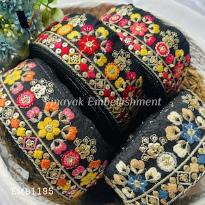 May include: A close-up of four rolls of black fabric trim with colorful embroidered floral designs and sequins. The trim is in a basket and the text "Vinayak Embellishment" and "EMB1195" are visible.
