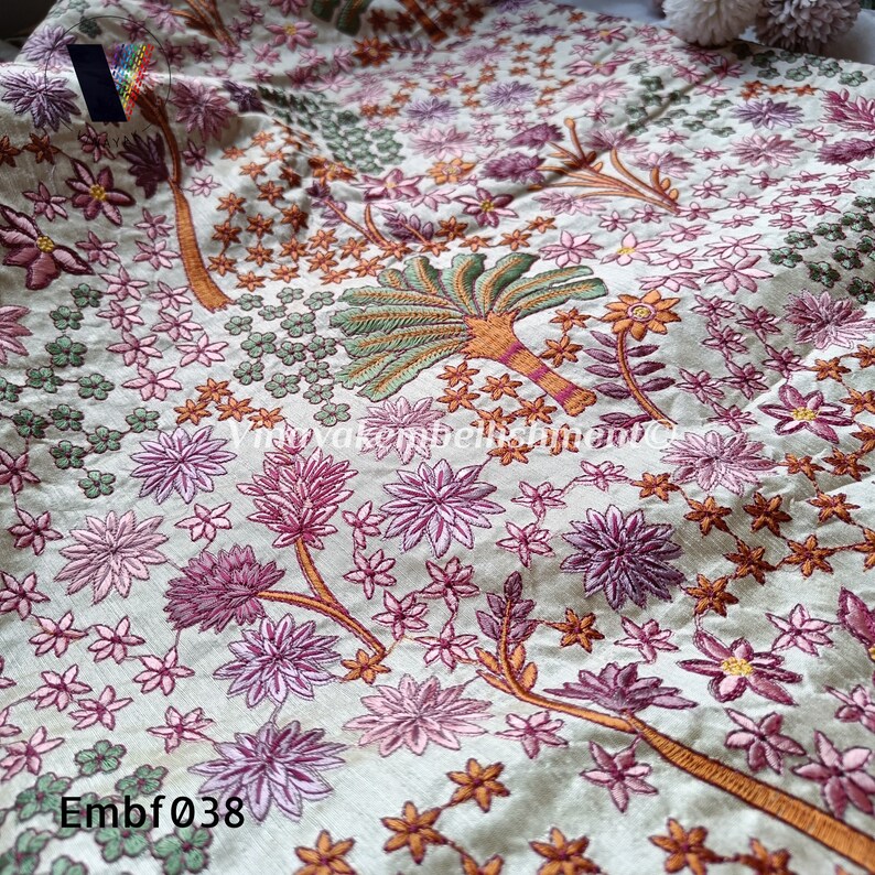May include: A close-up of a cream-coloured fabric with embroidered floral and tree designs in shades of pink, green, and orange. The fabric has a subtle sheen and a delicate texture. The text "Embf038" is visible in the bottom left corner of the image.