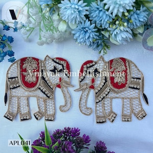 Könnte beinhalten: Zwei weiße bestickte Elefanten-Applikationen mit roten, goldenen und schwarzen Details. Die Elefanten sind in entgegengesetzte Richtungen gerichtet und haben eine perlenartige Verzierung an ihren Rüsseln. Der Text "APL041" ist in der linken unteren Ecke des Bildes sichtbar.