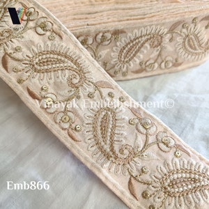 May include: A close-up of a pale pink embroidered trim with gold thread and sequins. The trim features a paisley design and floral motifs. The trim is folded and stacked, showing the intricate details of the embroidery.