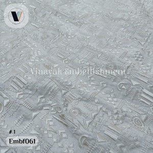 May include: White fabric with a geometric pattern embroidered in white thread. The fabric has a subtle sheen and a textured surface. The pattern is made up of a variety of shapes, including triangles, squares, and circles. The fabric is #1 Embf061.