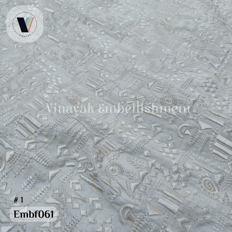 May include: White fabric with a geometric pattern embroidered in white thread. The fabric has a subtle sheen and a textured surface. The pattern is made up of a variety of shapes, including triangles, squares, and circles. The fabric is #1 Embf061.