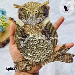 May include: A gold and brown owl applique featuring sequins and beads. The owl is perched on a branch with its wings outstretched. This applique is ideal for adding a touch of whimsy to any project.