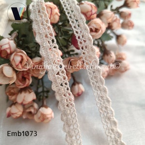 May include: Two pieces of off-white crochet trim with a delicate floral pattern. The trim is displayed against a background of pink and white flowers. The text "Emb1073" is visible in the lower left corner of the image.