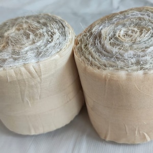 Könnte beinhalten: Zwei Rollen beigefarbener Stoff mit einer weißen, durchsichtigen, netzartigen Textur. Der Stoff ist fest aufgerollt und die Enden sind mit einem beigefarbenen Faden befestigt.
