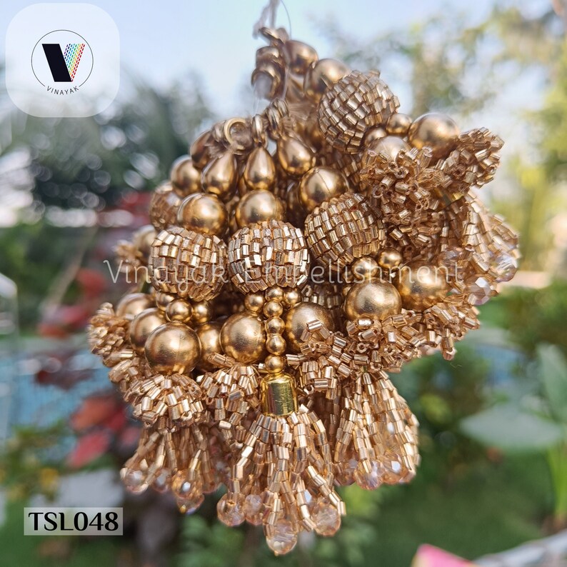 May include: A close-up of a bunch of gold-colored beaded tassels. The tassels are made of small, round beads and have a metallic sheen. The tassels are arranged in a cluster and are hanging from a string. The text "TSL048" is visible in the lower left corner of the image.