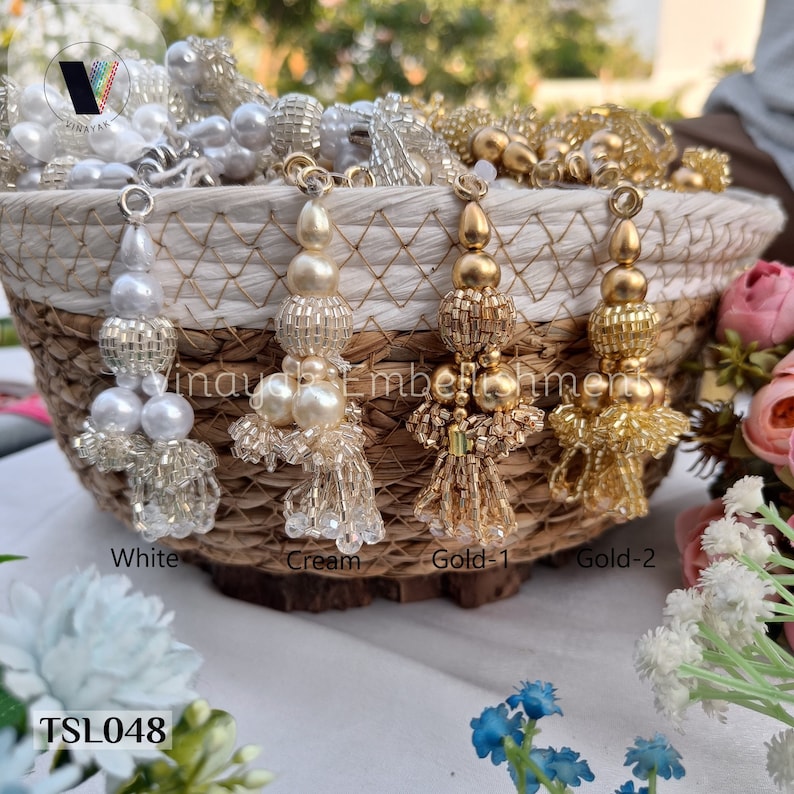 May include: Four decorative tassels in a wicker basket. The tassels are made of beads and have a fringe. The tassels are white, cream, gold, and gold. The text "TSL048" is visible at the bottom of the image.