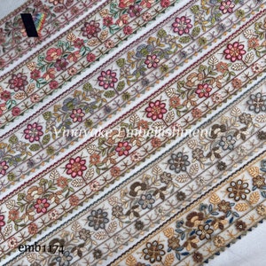 May include: A close-up of a white fabric with multiple rows of colorful embroidered floral trim. The trim features a variety of floral designs in shades of pink, green, blue, and brown. The text "Vinayake Embellishment" is visible in the center of the image.