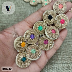 Puede incluir: Surtido de 10 botones redondos de tela bordada de color dorado con centros de diferentes colores. Los botones tienen un diseño en espiral y miden aproximadamente 2,5 cm de diámetro. Los colores de los centros incluyen naranja, morado, rosa, verde y negro.