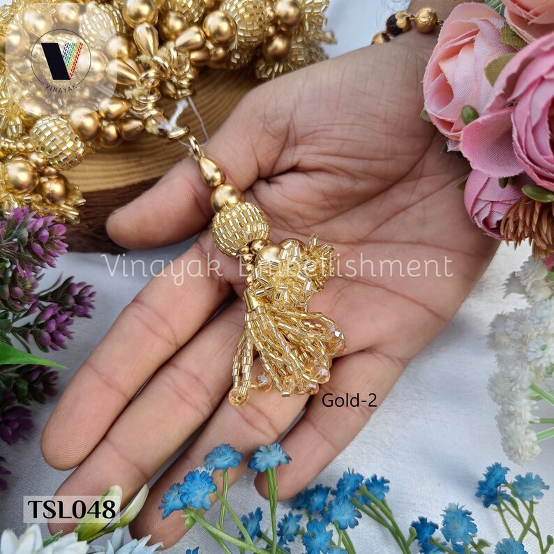 May include: Gold tassel trim with a beaded design. The tassel is made of small gold beads and has a round, beaded top. The trim is labeled "Gold-2" and "TSL048".