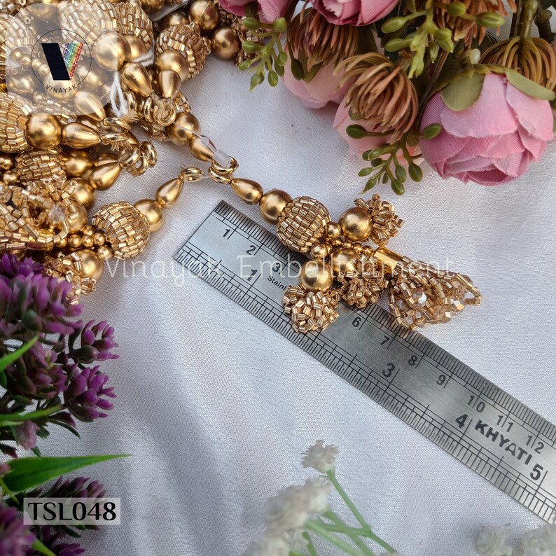 May include: Gold tassel trim with beads and a crystal accent. The trim is measured against a ruler showing it is approximately 6 inches long. TSL048