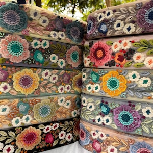 Puede incluir: Una colección de cintas de tela apiladas con intrincados bordados florales sobre malla transparente. Los diseños incluyen varias flores coloridas en tonos rosa, amarillo, azul y verde, con costuras detalladas y patrones de follaje.