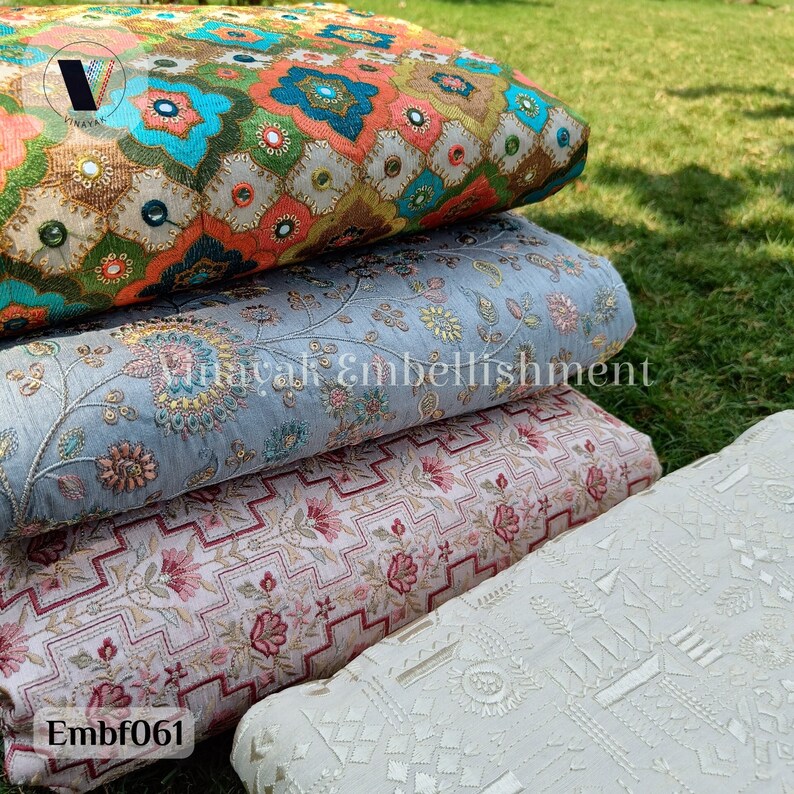 May include: Four different fabric swatches stacked on top of each other. The top swatch is a colorful floral pattern with sequins and mirror embellishments. The second swatch is a light blue fabric with a floral pattern. The third swatch is a pink and white fabric with a geometric pattern. The bottom swatch is a white fabric with a geometric pattern. The text "Embf061" is visible on the bottom swatch.