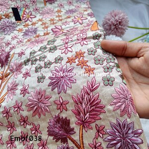 May include: A close-up of a cream-coloured fabric with pink, green and orange embroidered flowers. The fabric has the text "Embf038" in the bottom left corner.