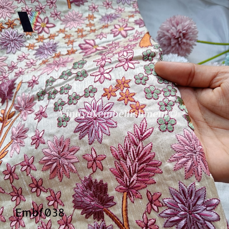 May include: A close-up of a cream-coloured fabric with pink, green and orange embroidered flowers. The fabric has the text "Embf038" in the bottom left corner.