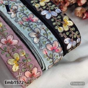 Op de afbeelding: Close-up van geborduurde bloemenrand in tinten paars, blauw en zwart. De rand heeft gedetailleerde bloempatronen met roze, gele en witte accenten. De tekst "Emb1572" is zichtbaar linksonder.