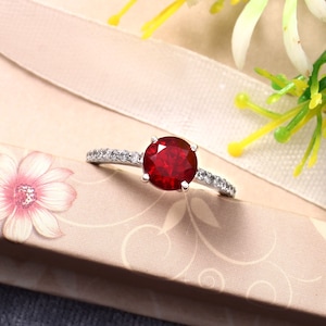 May include: A silver-toned ring featuring a large, round, deep red gemstone set in a four-prong mount. The band is accented with a row of small, clear, sparkling stones embedded along both sides of the central setting, creating a bright contrast.