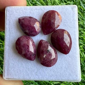 Puede incluir: Cinco gemas facetadas, en forma de lágrima, de color rojo intenso, dispuestas sobre una superficie blanca. Las gemas tienen una superficie pulida y reflectante, sobre un fondo de hierba verde.