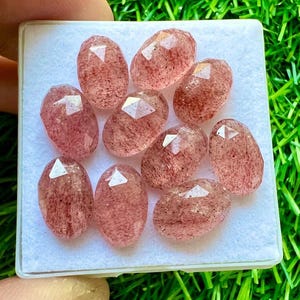 Könnte beinhalten: Eine Sammlung von zehn ovalen, facettierten, rosafarbenen Quarzsteinen. Die Steine sind durchscheinend und haben eine rotbraune Farbe.