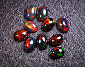 May include: A collection of ten oval-shaped black opals with iridescent red, green, and blue flashes. The opals are arranged on a textured brown surface.