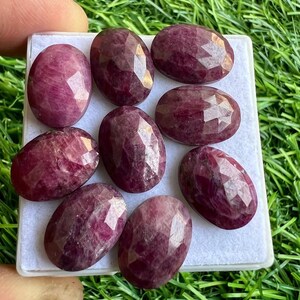May include: Eight oval-shaped, faceted ruby gemstones in various shades of pink and red. The gemstones are displayed on a white surface.
