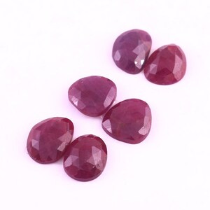 Puede incluir: Seis piedras preciosas de rubí facetadas en forma de corazón, de color rojo intenso. Las piedras están dispuestas en una fila sobre un fondo blanco.