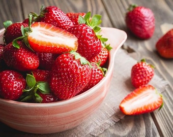 May include: A bowl filled with ripe, red strawberries, some halved to reveal their juicy interior. Green leaves peek out from the fruit. Several whole strawberries are scattered on a wooden surface, alongside a linen cloth.