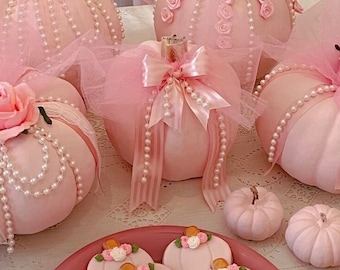 Peut inclure: Plusieurs citrouilles décoratives roses ornées de perles, de tulle et de nœuds. Certaines citrouilles présentent des détails de roses. Une assiette de biscuits en forme de citrouille, décorés de fleurs, complète l'ensemble, créant un thème cohérent.
