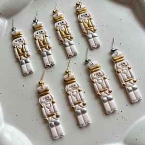 Op de afbeelding: Acht witte en gouden notenkraker oorbellen met zilveren en gouden accenten, gepresenteerd op een wit bord met geschulpte rand. Elke oorbel heeft een gedetailleerd notenkraker ontwerp.
