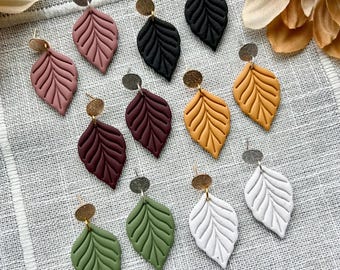 May include: A collection of leaf-shaped earrings in various colors, including pink, black, burgundy, yellow, green, and white. Each earring features a textured leaf design and a small gold-colored disc at the top. The earrings are displayed on a textured, light gray surface.