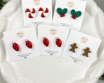 May include: A collection of Christmas-themed stud earrings displayed on white cards. Designs include red and white peppermint candies, green holly with red berries, red Christmas lights, and gingerbread men. The brand name and social media handles are printed on the cards.