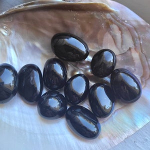 May include: A collection of polished black obsidian stones, oval-shaped, resting inside a large, iridescent seashell. The stones have a glossy, reflective surface, and the shell displays a range of colors.