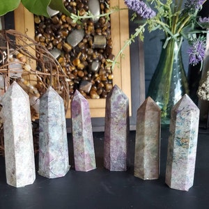 May include: Six polished, multi-faceted, pink and green gemstone points, also known as wands or towers, arranged in a row on a black surface.