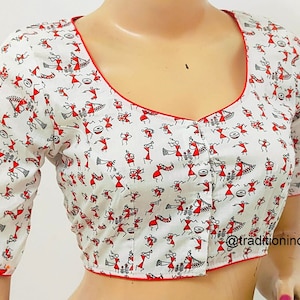 May include: White cotton blouse with a red trim and a pattern of stick figures in red and black. The stick figures are dancing and playing instruments. The blouse has a scoop neck and short sleeves. The text "@traditionindia" is visible in the bottom right corner of the image.