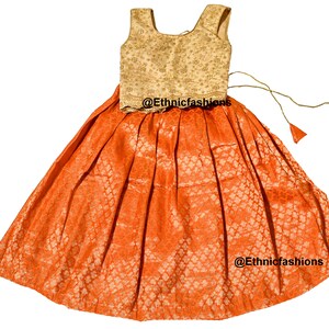May include: A two-piece outfit for a young girl, featuring a sleeveless, gold-colored top with a floral pattern and an orange skirt with a matching floral pattern. The skirt has a drawstring closure and is pleated. The outfit is decorated with gold embroidery and the text "@Ethnicfashions" appears on the skirt.