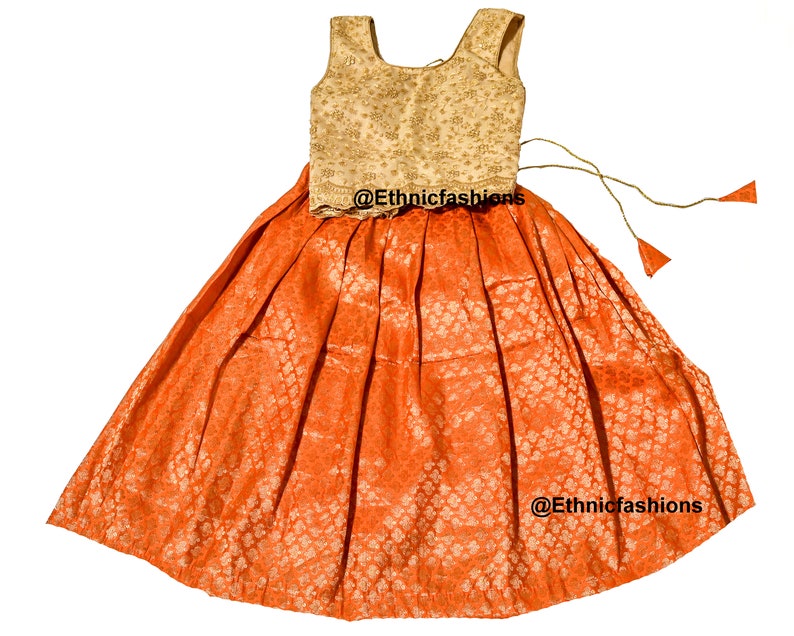 May include: A two-piece outfit for a young girl, featuring a sleeveless, gold-colored top with a floral pattern and an orange skirt with a matching floral pattern. The skirt has a drawstring closure and is pleated. The outfit is decorated with gold embroidery and the text "@Ethnicfashions" appears on the skirt.
