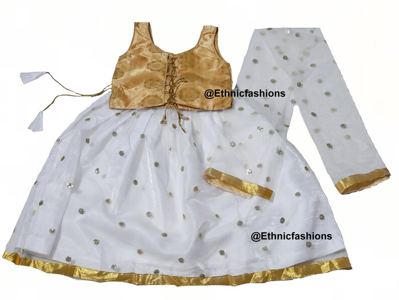 May include: A white and gold girls' lehenga choli with a gold embroidered bodice and a white skirt with sequins. The skirt has a gold trim and the set includes a white dupatta with gold trim. The text '@Ethnicfashions' appears twice on the image.