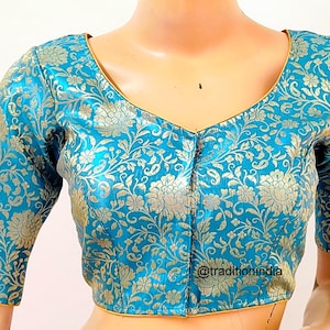 May include: A teal blue brocade blouse with a gold floral pattern and a V-neckline. The blouse has short sleeves and a hook and eye closure at the front. The text "@traditionindia" is visible in the bottom right corner of the image.