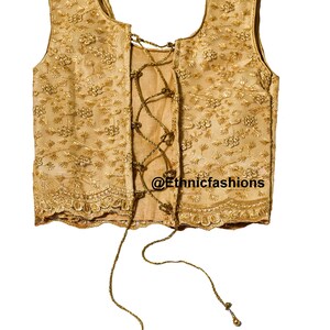 May include: A gold sleeveless blouse with a lace-up back and floral embroidery. The blouse has a scalloped hem and is tied with a gold cord.