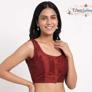 May include: A sleeveless blouse in a deep maroon color with a subtle textured finish. The garment features a scoop neckline and a front button placket that runs vertically down the center. The fabric has a slight sheen, and the design is simple.