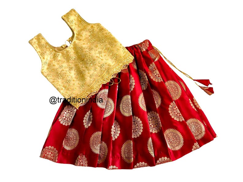 May include: A red and gold girls' outfit with a sleeveless, embroidered top and a skirt with a circular pattern. The skirt has a gold tassel.